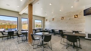 Een ruime eetruimte met meerdere tafels en stoelen, grote ramen met uitzicht op de bergen en een houten wand met ingebouwde verlichting.