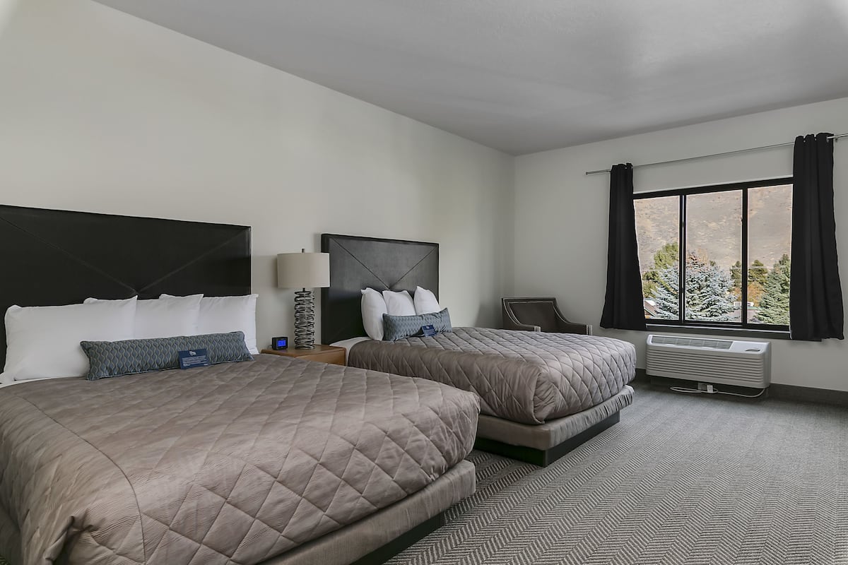 A hotel room with two beds, a desk, a chair, a window with curtains, and an air conditioning unit.