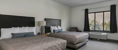 A hotel room with two beds, a desk, a chair, a window with curtains, and an air conditioning unit.