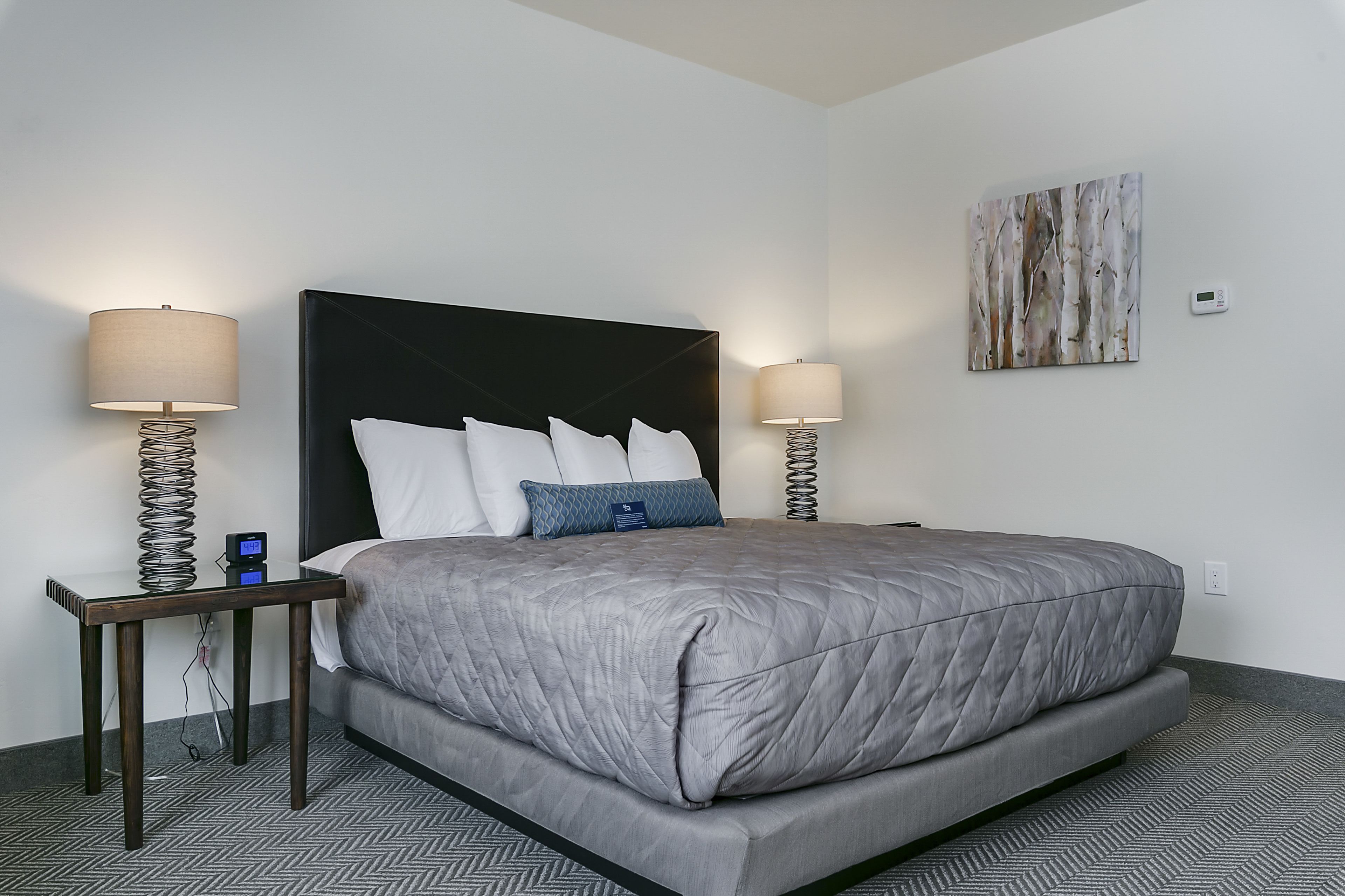 Una habitación de hotel moderna con una cama grande, dos mesitas de noche con lámparas, un termostato en la pared y una obra de arte abstracta.