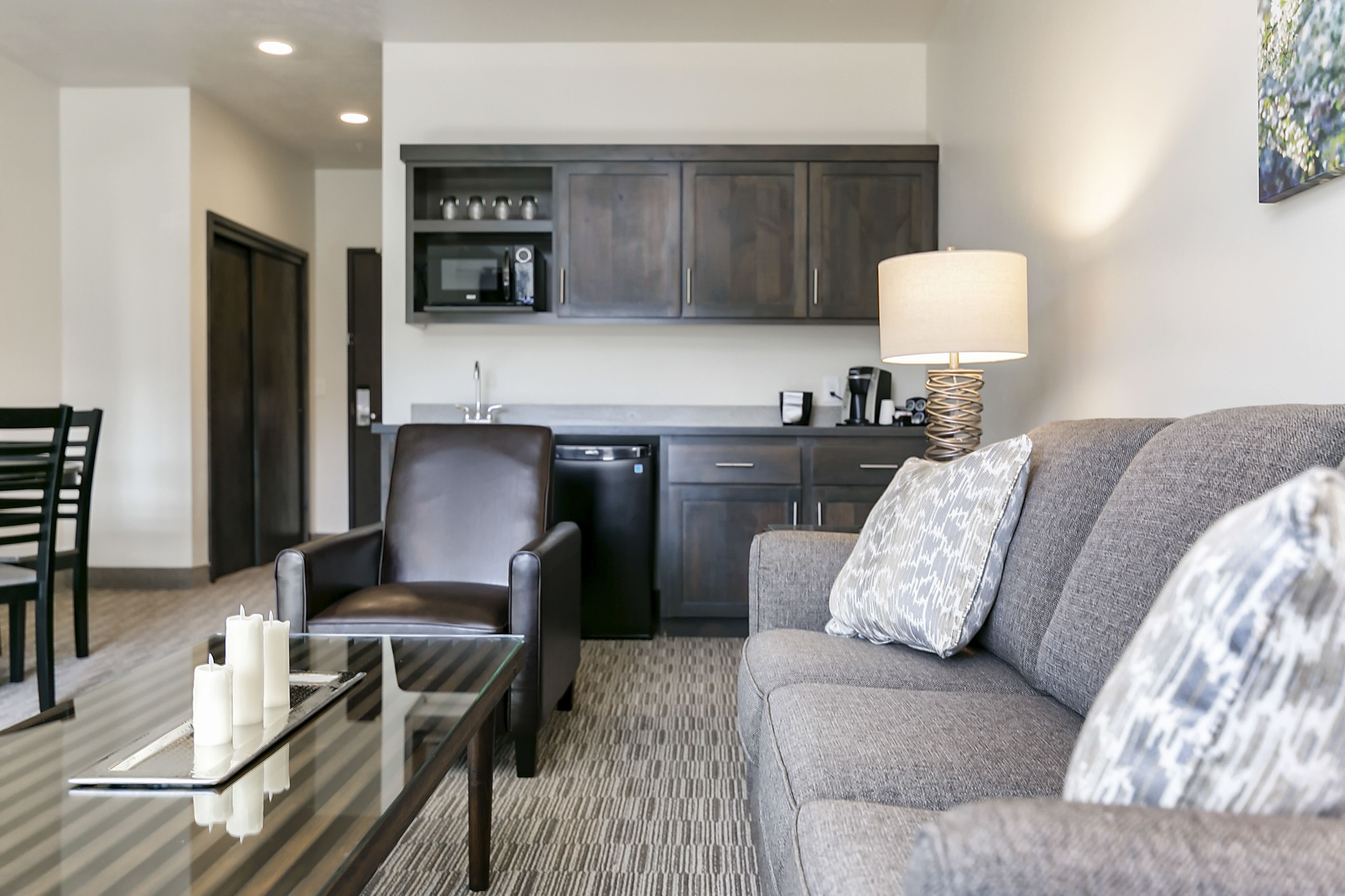 A modern living room with a grey sofa, a black leather chair, a glass coffee table, and a kitchen area in the background.