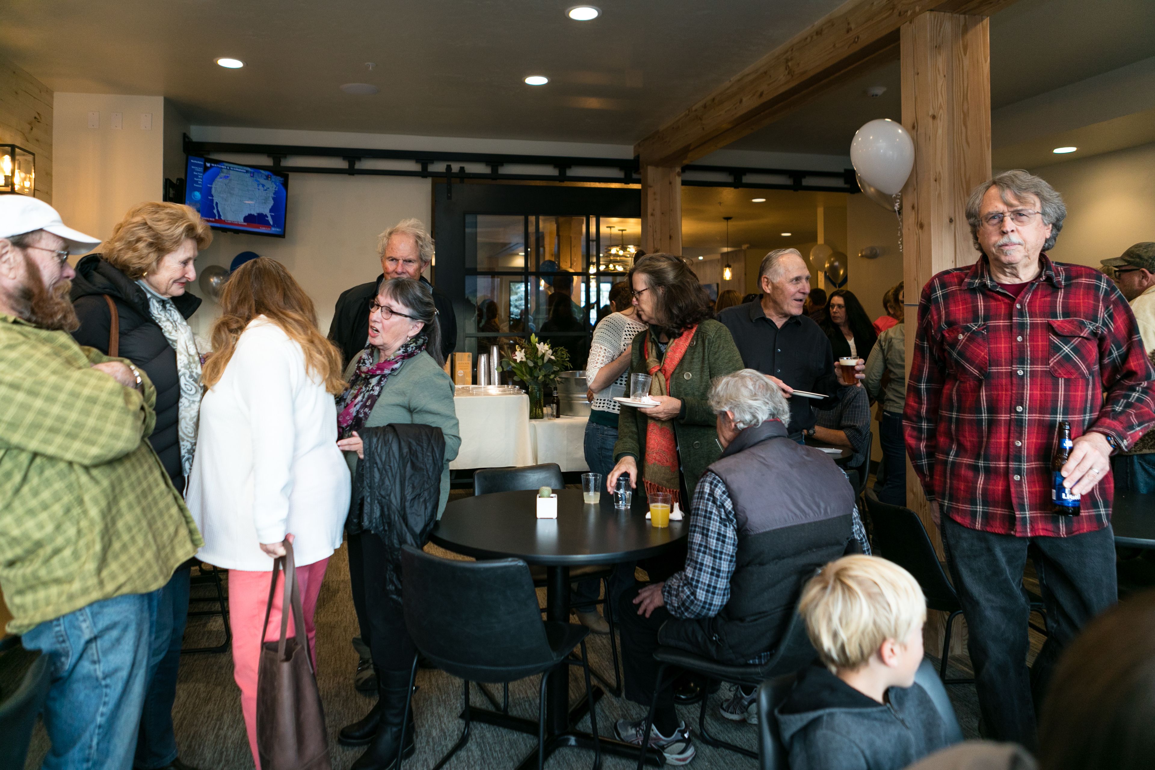 A group of people in a social setting with tables and chairs, a television mounted on the wall, and a wooden ceiling structure.