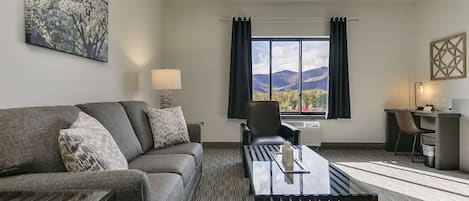 A hotel room with a sofa, a table, a chair, a desk, and a large window with a mountain view.