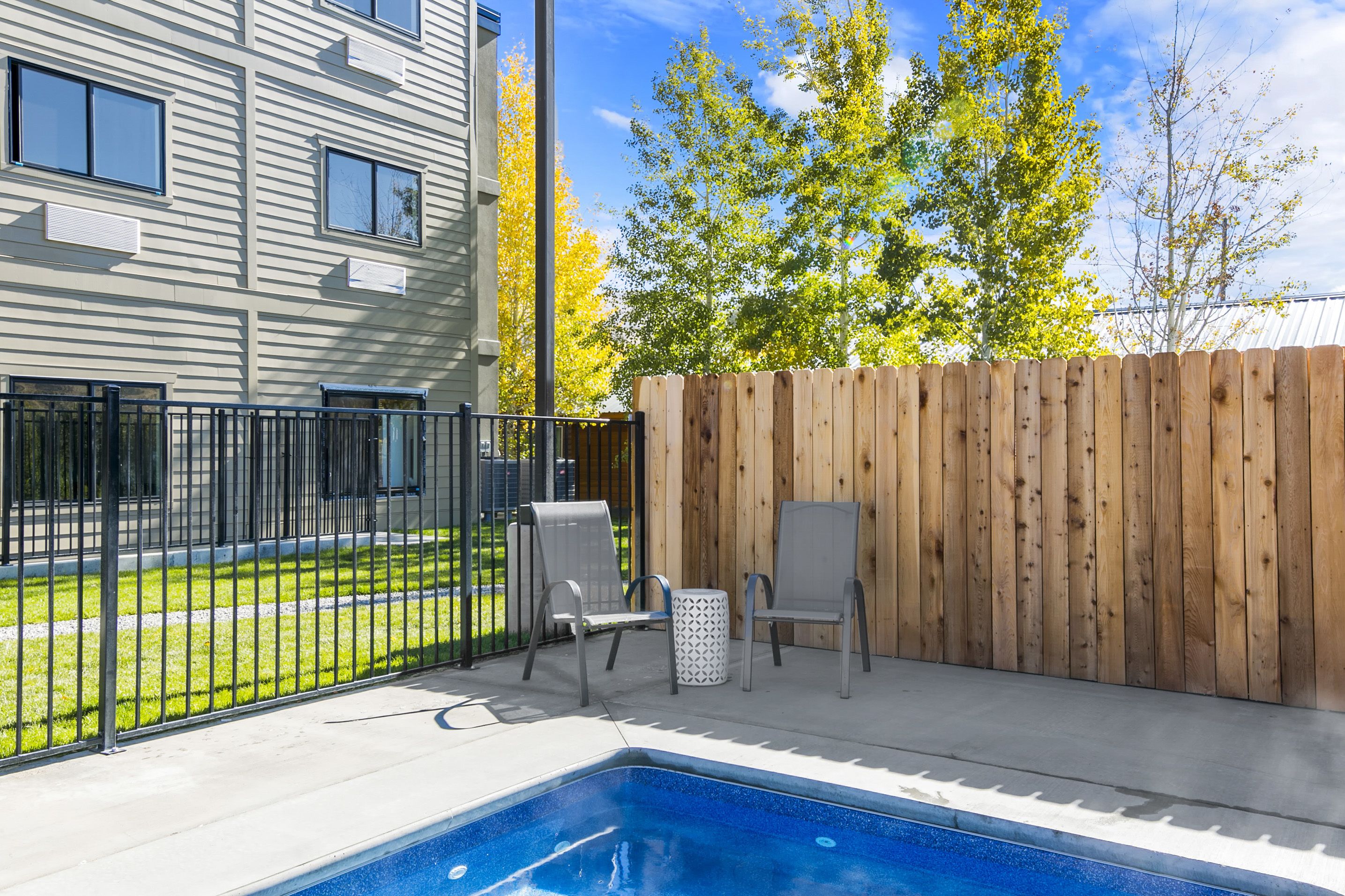 A backyard with a swimming pool, two chairs, a wooden fence, and a building with windows.