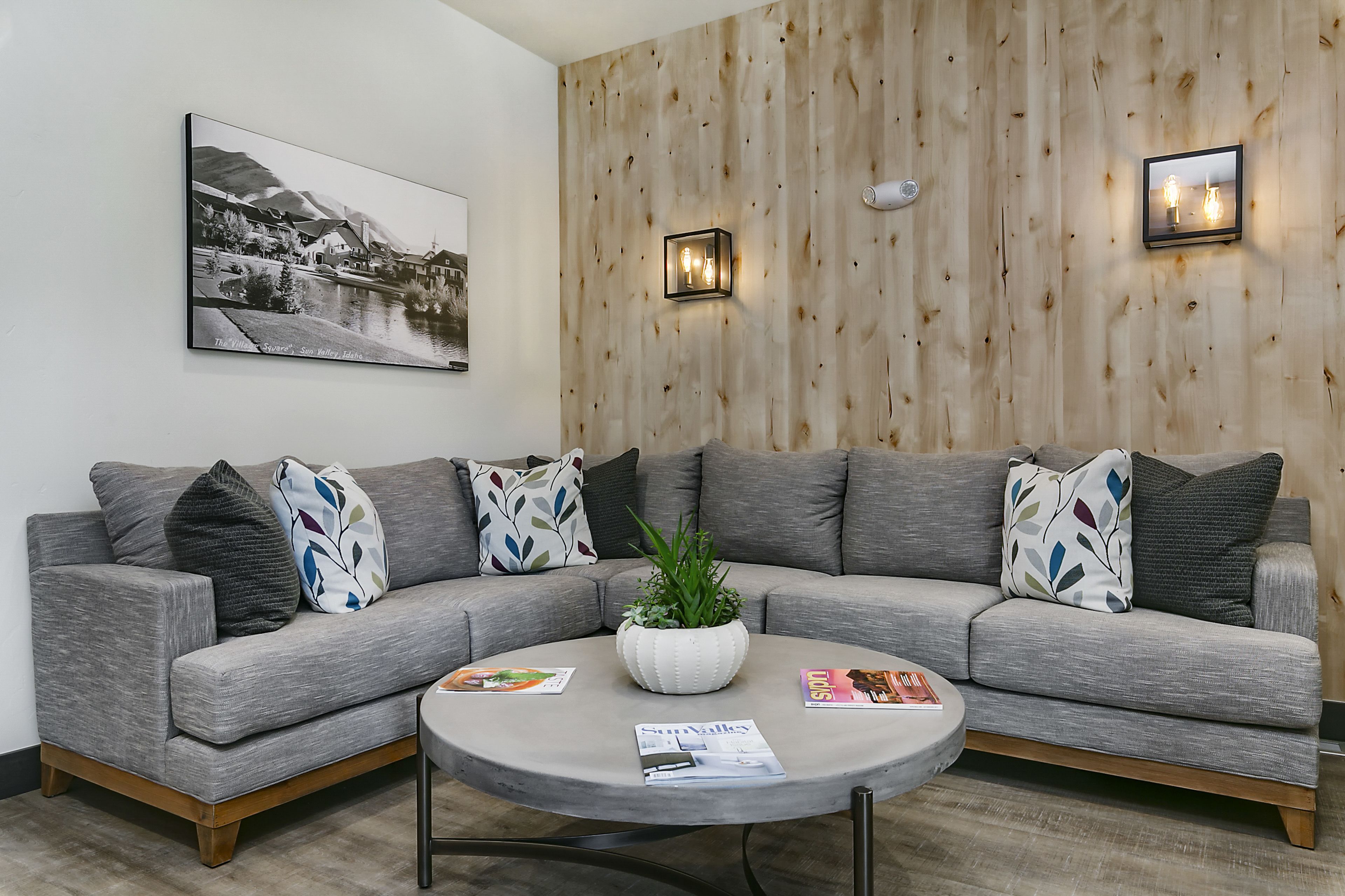 En moderne stue med en grå modulsofa, et rundt sofabord og en potteplante. Der er et sort-hvidt billede i ramme på væggen og to vægmonterede lamper.
