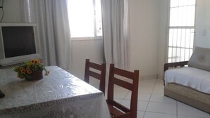 A room with a bed, a table with a lace tablecloth, a TV, and a window with curtains. - Férias na Beira da Praia em Cabo Frio (Cabo Frio)