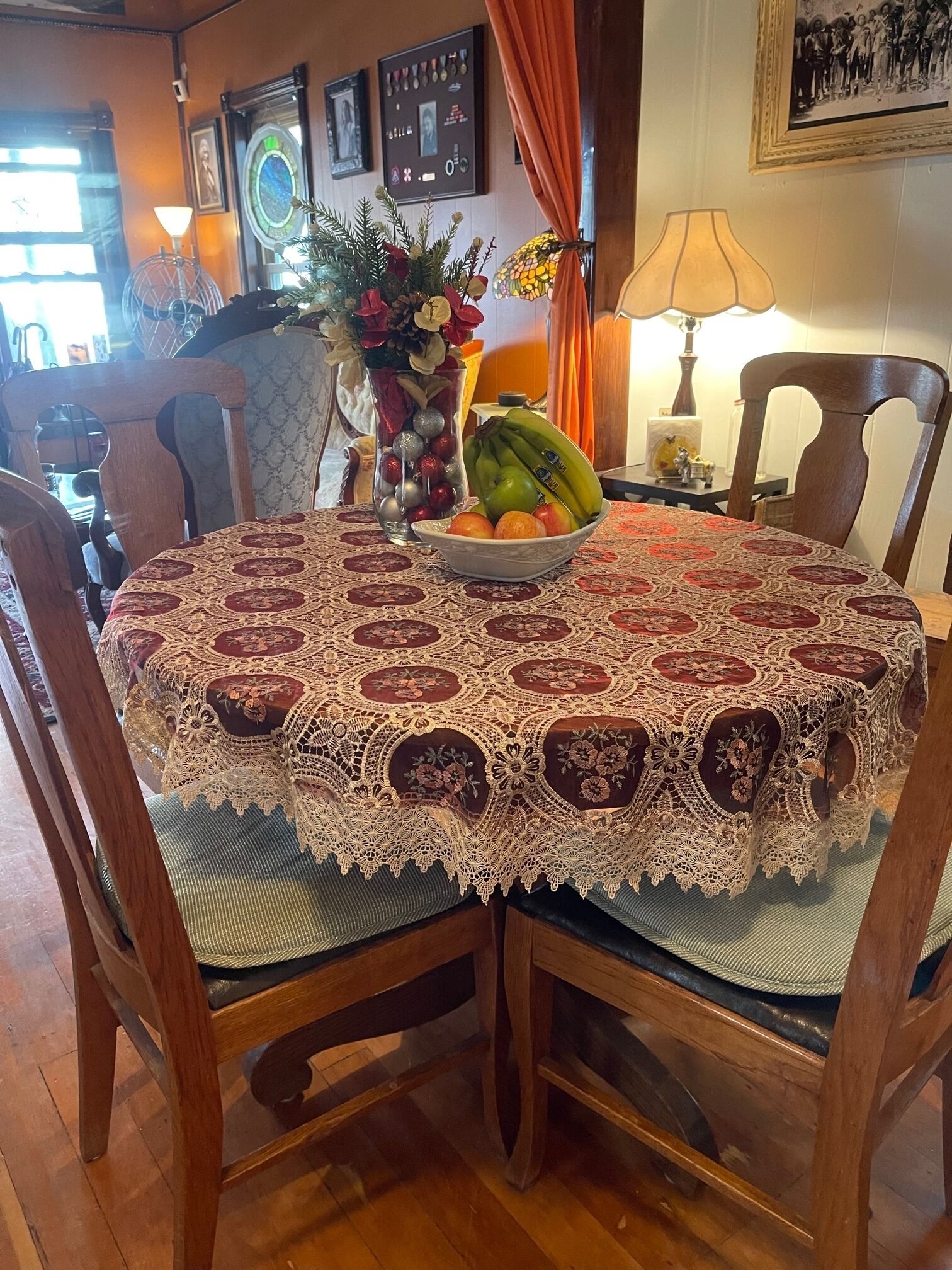 A dining table with a lace tablecloth, surrounded by wooden chairs. A bowl of fruit and a vase with flowers are on the table.