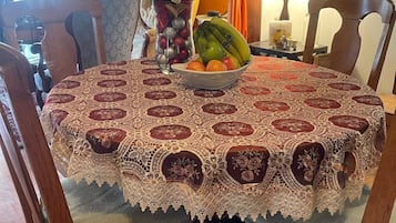A dining table with a lace tablecloth, surrounded by wooden chairs. A bowl of fruit and a vase with flowers are on the table.
