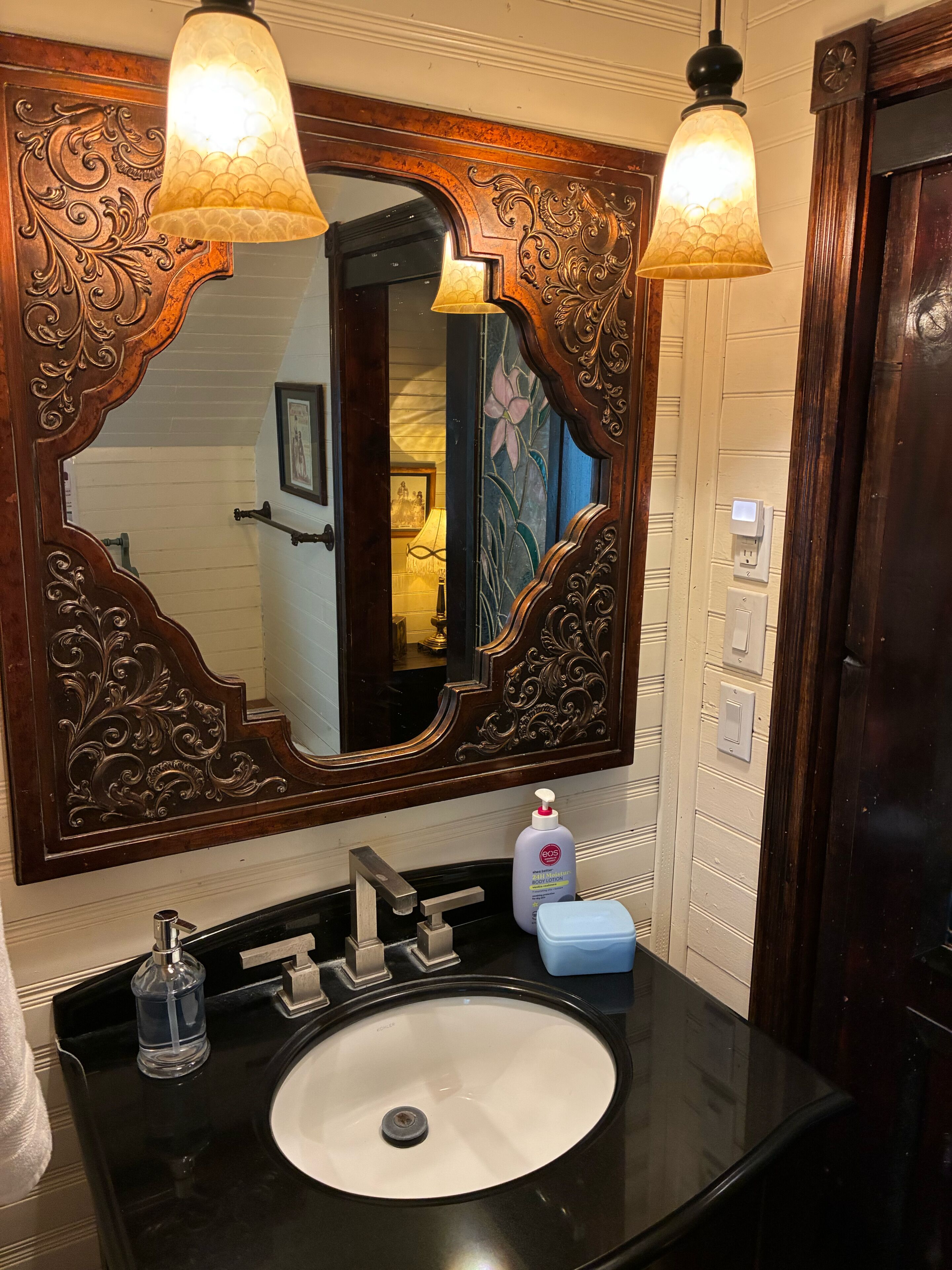 Un baño con un espejo ornamentado, un lavabo negro y dos luces fijadas a la pared.