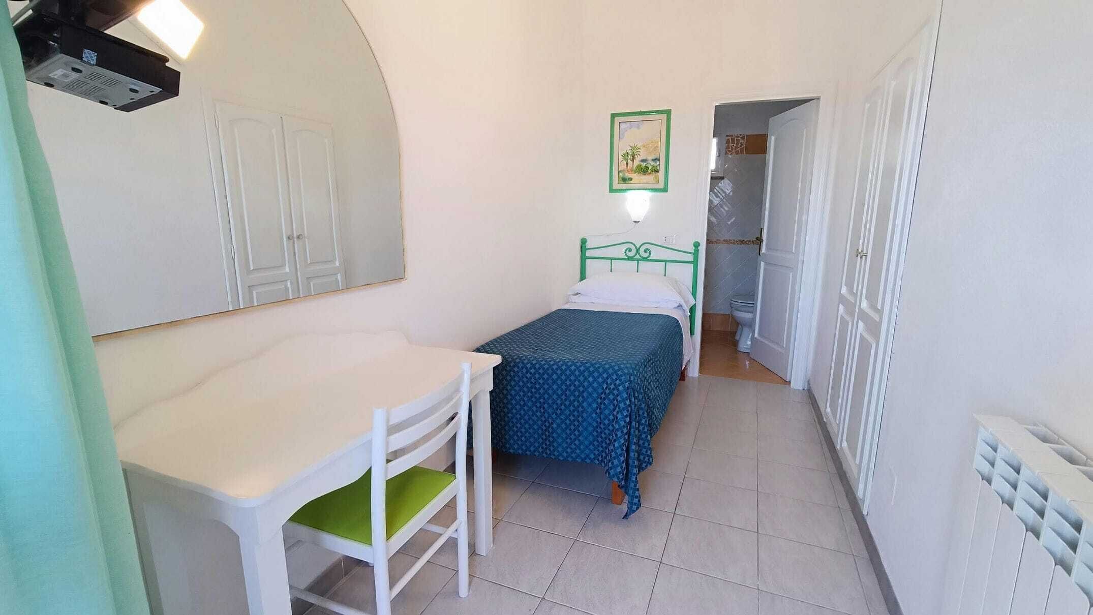 Una habitación pequeña y estrecha con una cama individual, un escritorio y una silla. Se observa un radiador a la derecha y una puerta que da a otra habitación.