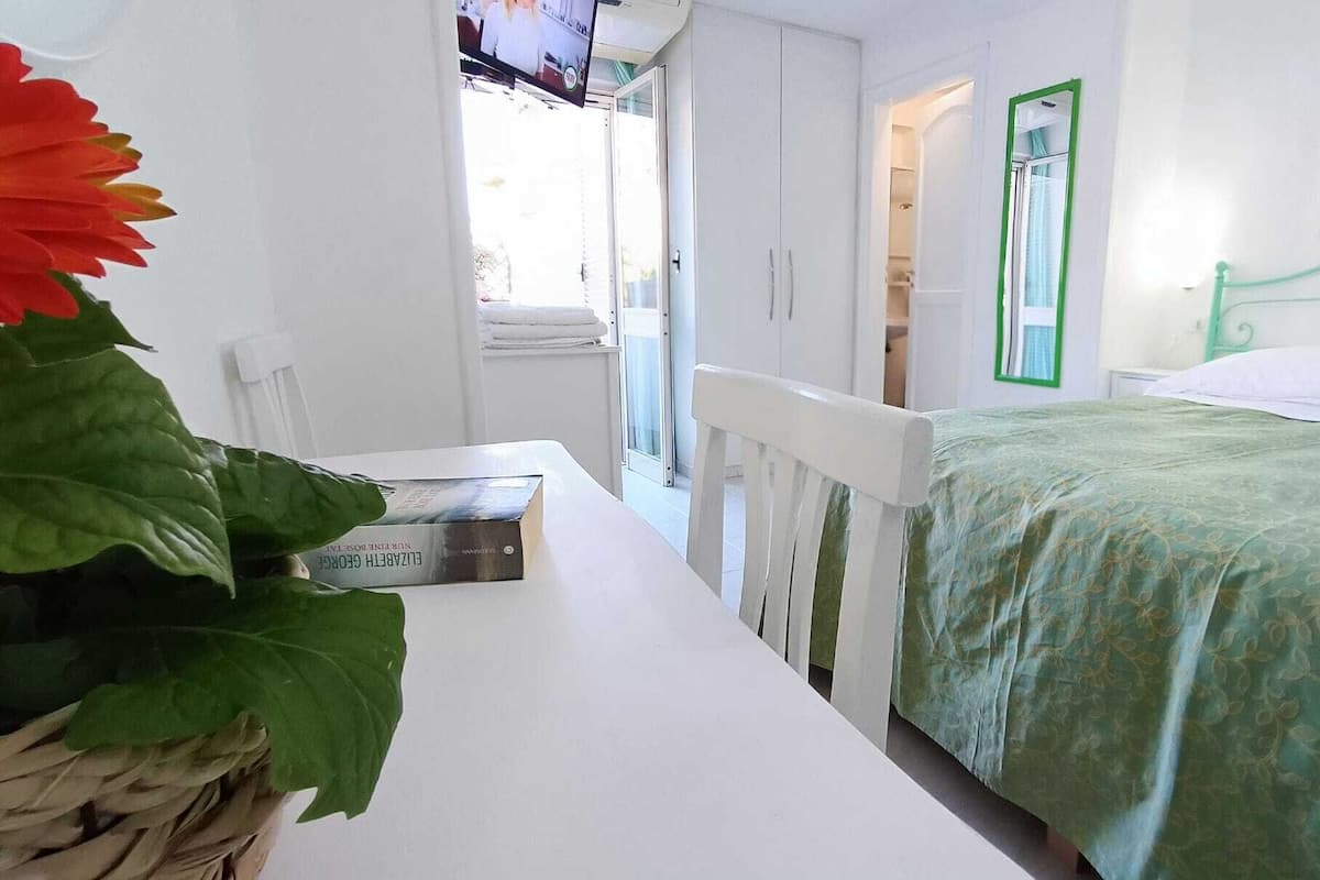 Een slaapkamer met een bed, een nachtkastje, een raam met groene omlijsting en een wand met een bloemschilderij.