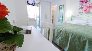 A bedroom with a bed, a nightstand, a window with a green frame, and a wall with a floral painting. - Hotel Casa Nicola (Serrara Fontana)