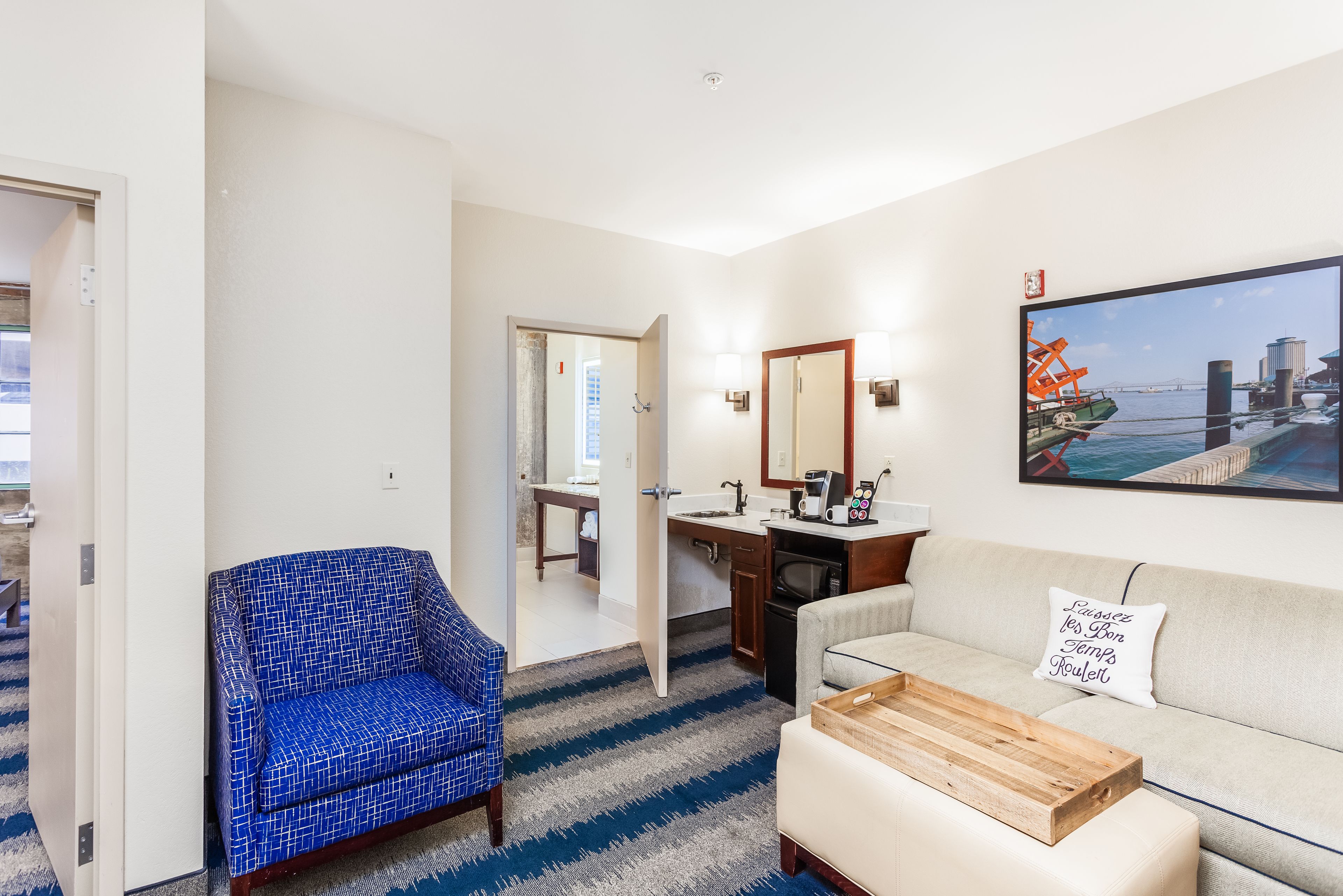 A hotel room with a blue patterned armchair, a beige sofa, a wooden coffee table, a kitchen area with a sink and stove, and a large window with a cityscape view.