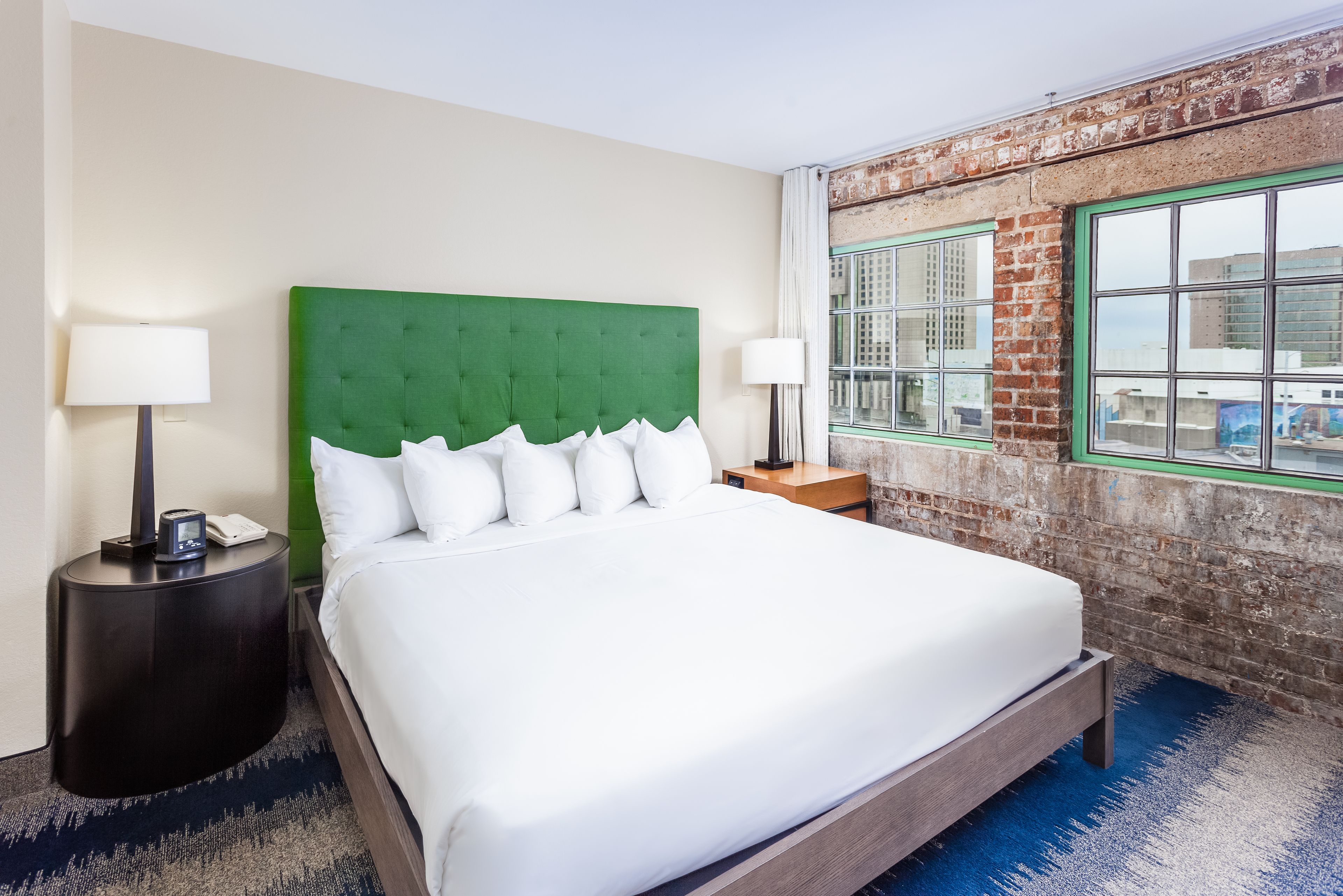 A hotel room with a large bed, green headboard, white bedding, a nightstand with a lamp, and a view of a brick wall and window.