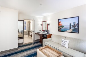 Ein modernes Hotelzimmer mit einer Küchenzeile, einem Sofa mit einem Zierkissen, einem großen Fenster mit Blick auf die Stadtlandschaft und einem blau-weiß gestreiften Teppich.