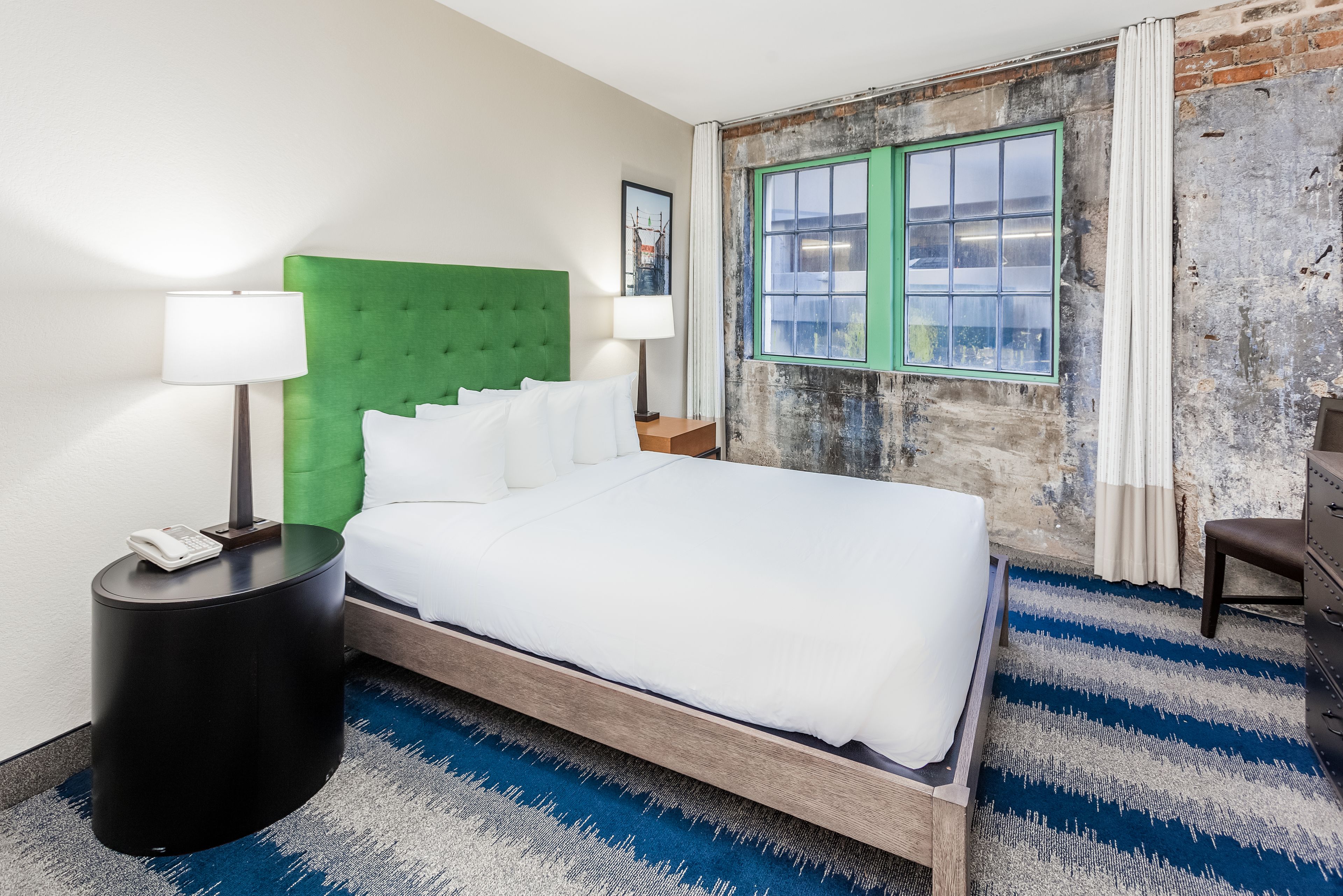 A hotel room with a green upholstered headboard, a large bed with white bedding, a round black side table with a lamp, a blue and white striped rug, and a view of a city through a green-framed window.