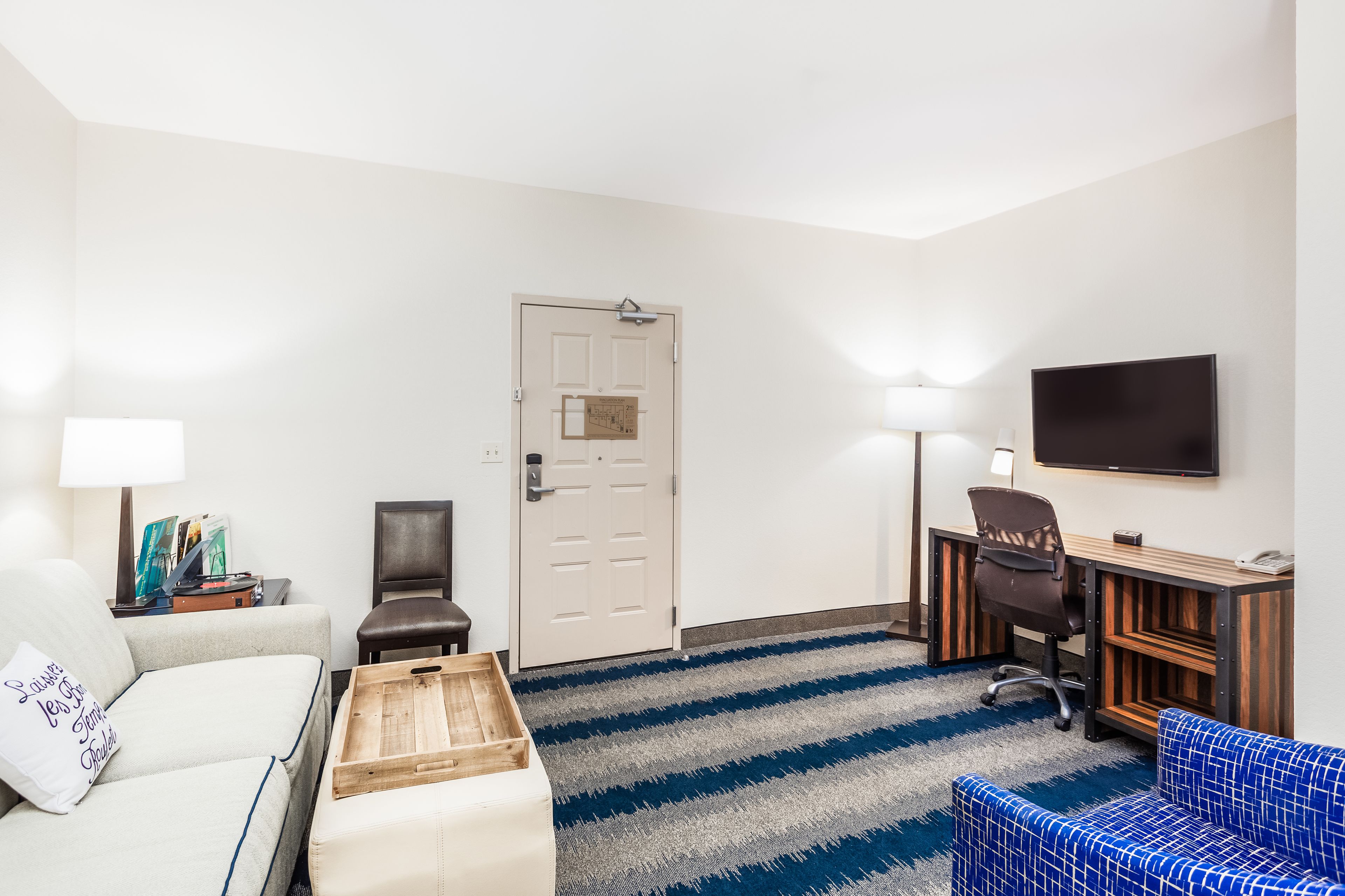 A hotel room with a sofa, a small wooden table, a blue patterned chair, a desk with a chair, a television, and a door with a light switch.
