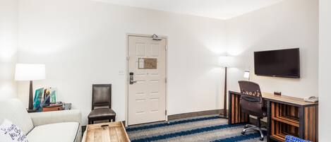 A hotel room with a sofa, a small wooden table, a blue patterned chair, a desk with a chair, a television, and a door with a light switch.