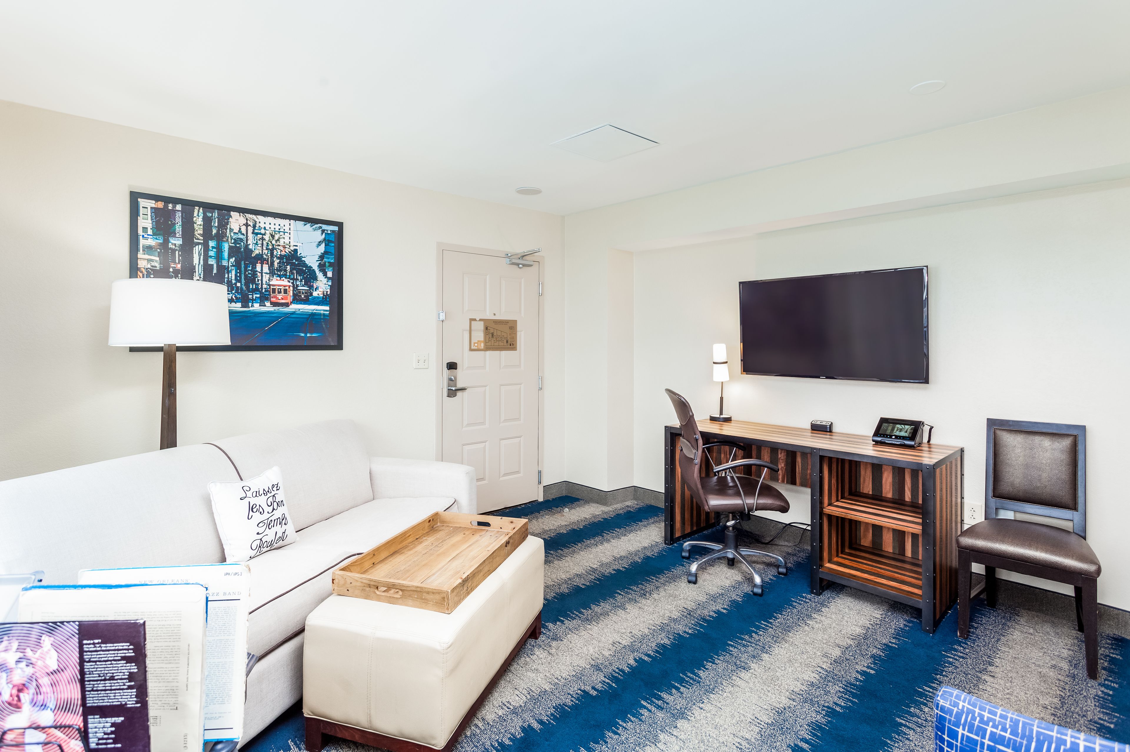 Habitación de hotel con sofá blanco, mesa de centro de madera, escritorio con computadora, silla, televisor y alfombra azul estampada.