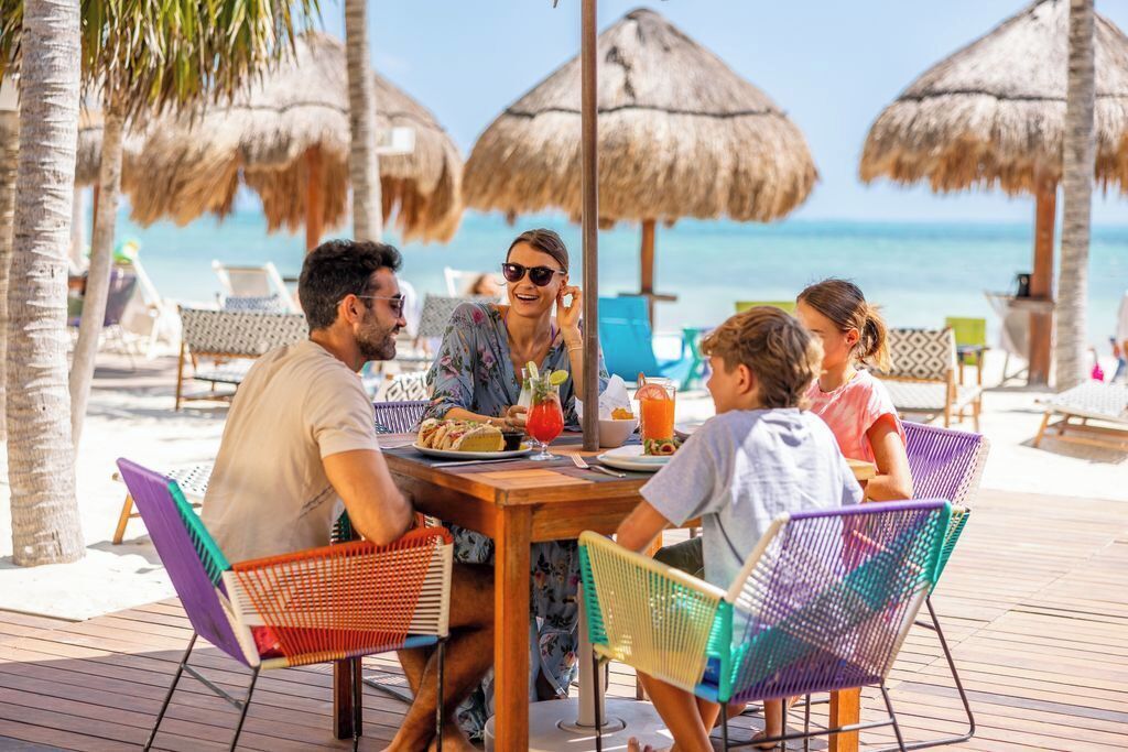 A group of people dining at a wooden table with colorful chairs on a deck, with a beach and ocean in the background.