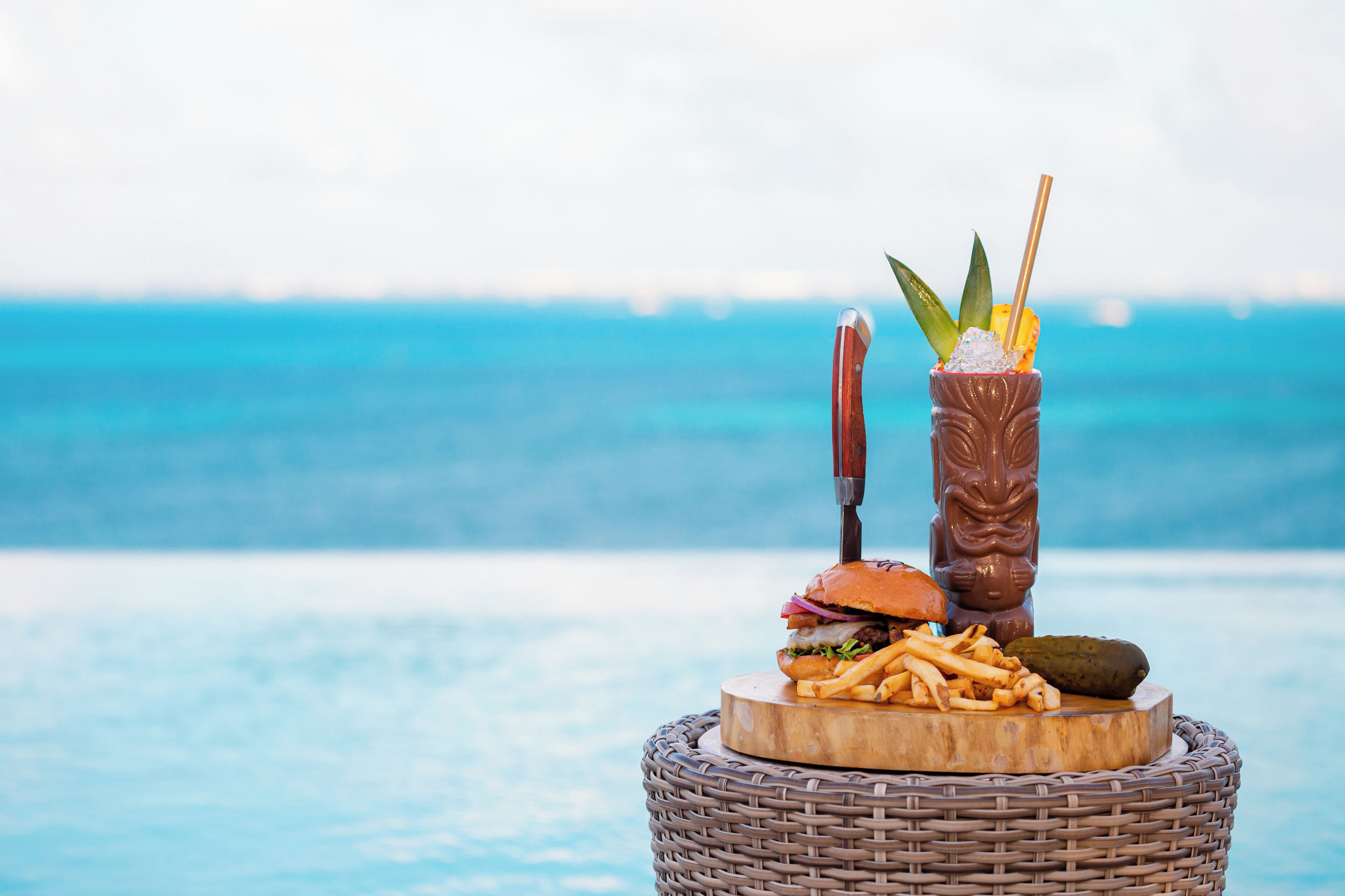 A wicker table with a burger, fries, a cocktail, and a knife and fork.