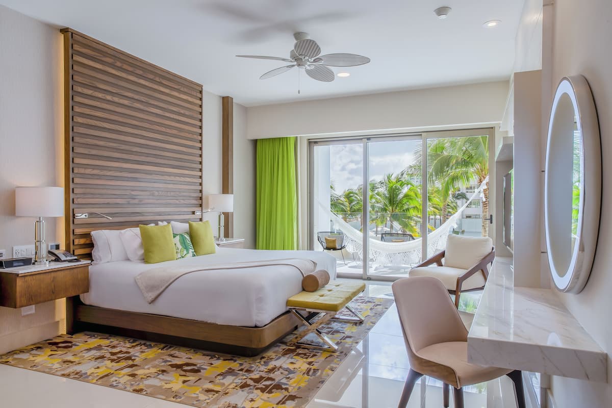 Een moderne hotelkamer met een groot bed, een bureau, een stoel, een plafondventilator en uitzicht op een hangmat en palmbomen buiten.