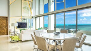 A modern dining area with a large glass table and chairs, a television, and a view of the ocean.