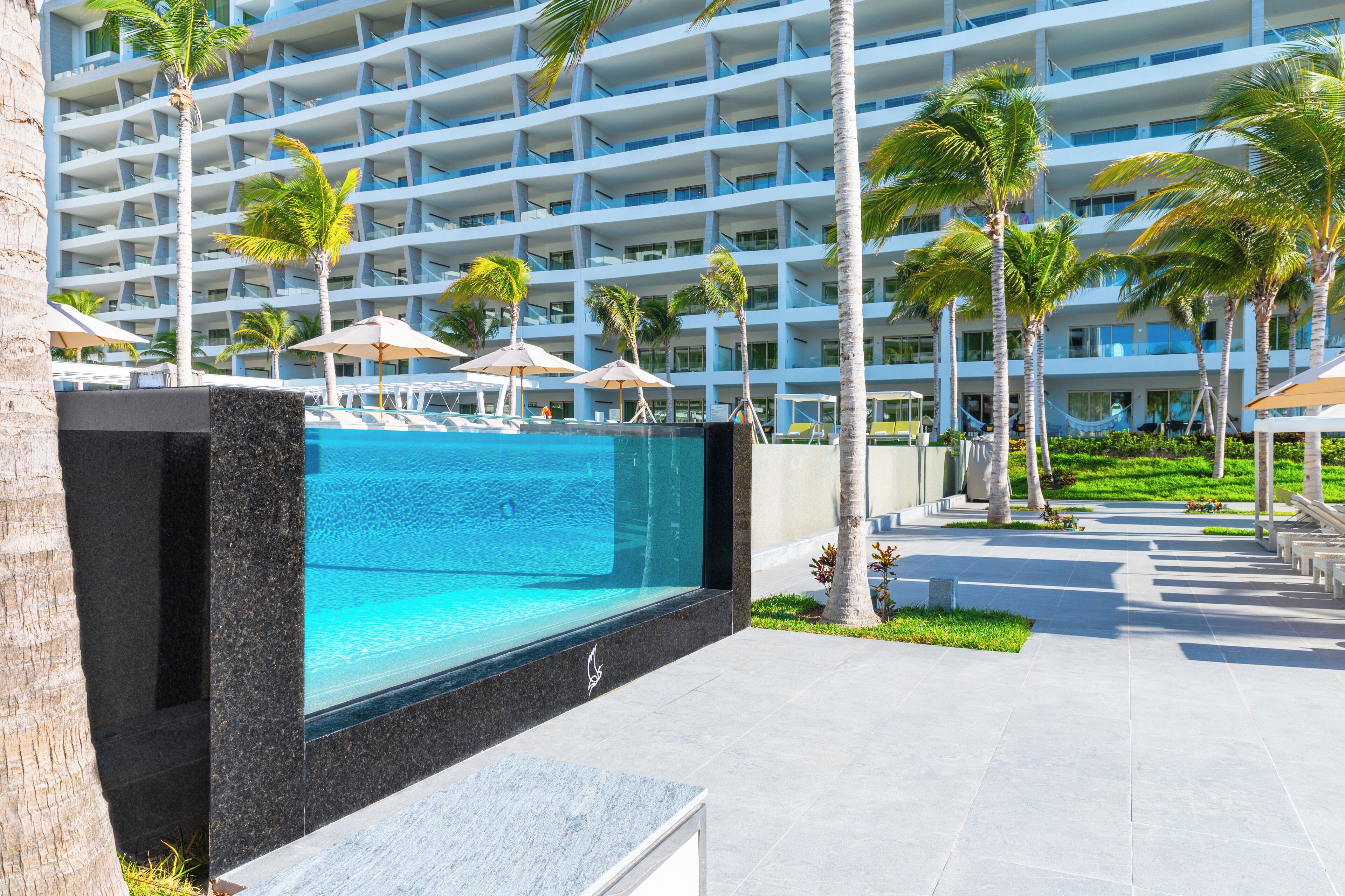 A modern hotel with a large pool, palm trees, and a multi-story building with balconies.