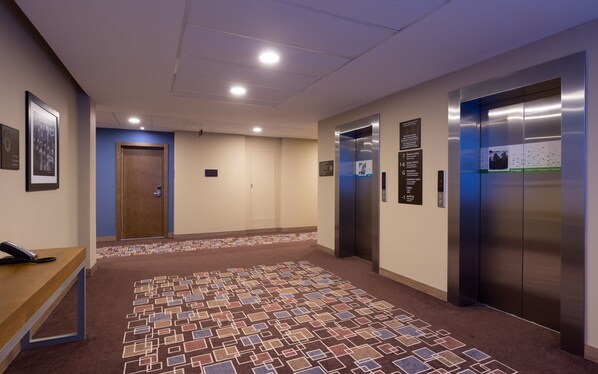Vestíbulo de hotel con una recepción, dos ascensores y una alfombra estampada.