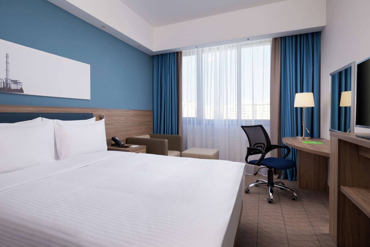 Une chambre d’hôtel avec un grand lit, un bureau avec une chaise, une petite table, une lampe et une fenêtre avec des rideaux.