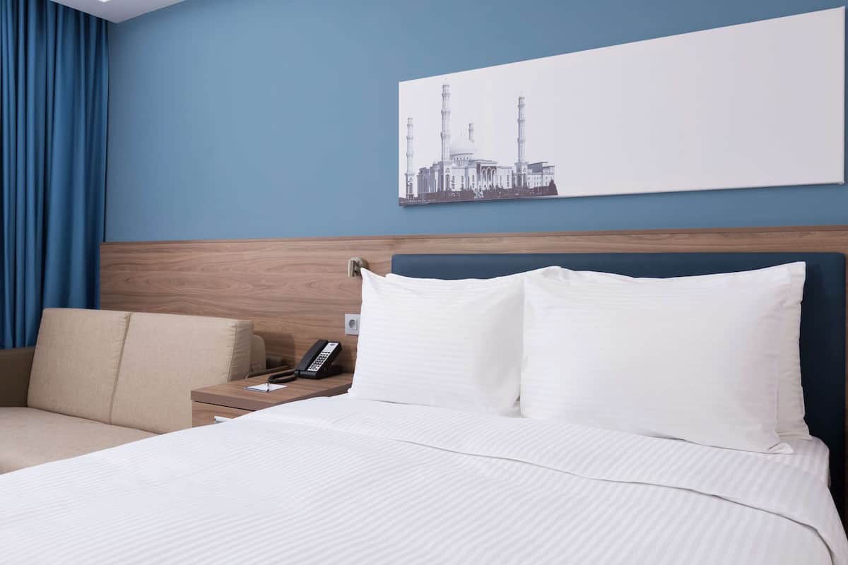 Une chambre d’hôtel avec un grand lit, une table de chevet avec un téléphone et un tableau représentant un bâtiment accroché au mur.