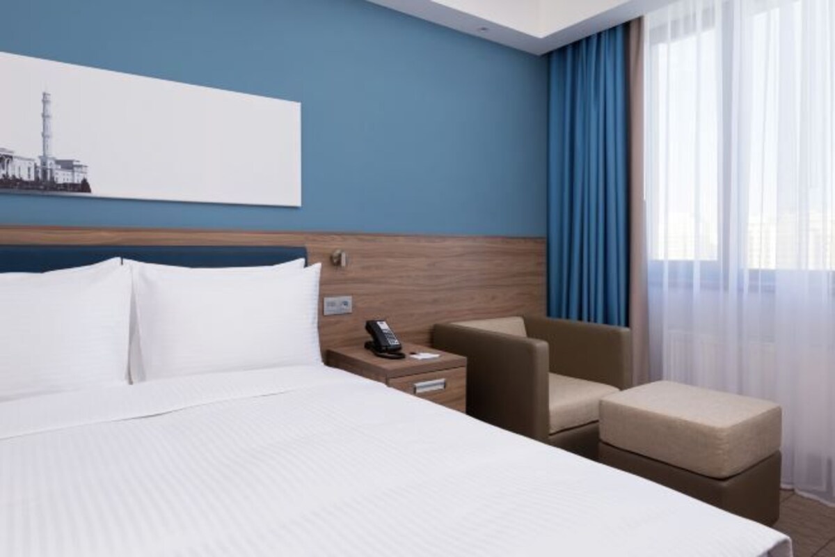 A hotel room with a large bed, a bedside table with a phone, a chair, and a small ottoman.