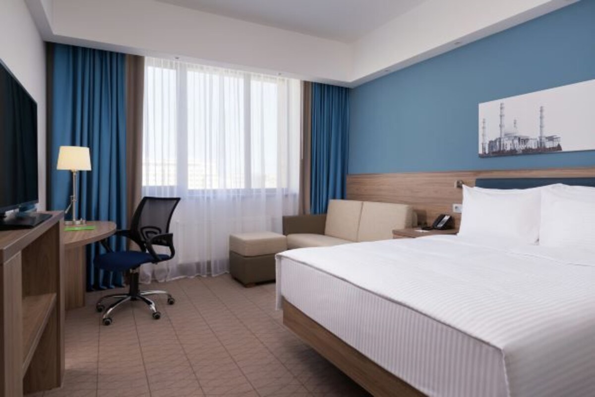 A hotel room with a bed, a desk with a computer, a chair, a small table, and a window with curtains.