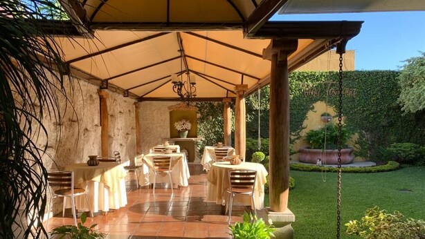 An outdoor patio with tables and chairs, a hanging light fixture, and a garden with a hedge and potted plants. - Hostal Villa Toscana (Guatemala City)
