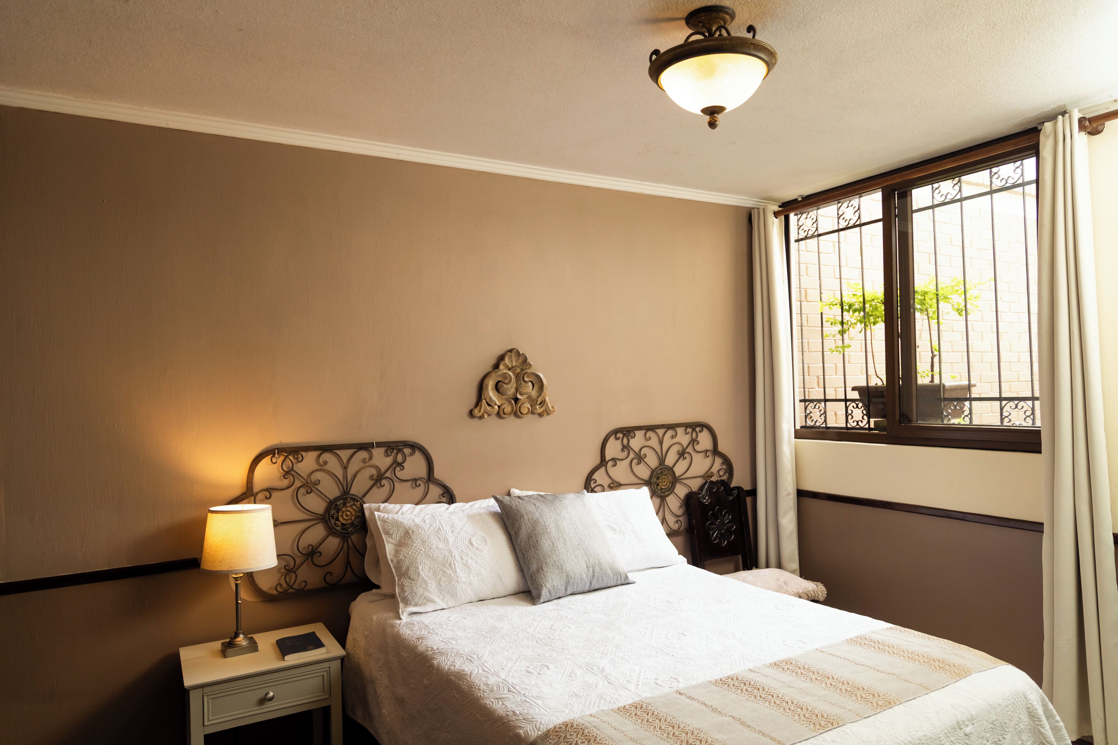 Hotelkamer met een bed, nachtkastje, lamp, decoratief wandobject en een raam met uitzicht op groen.