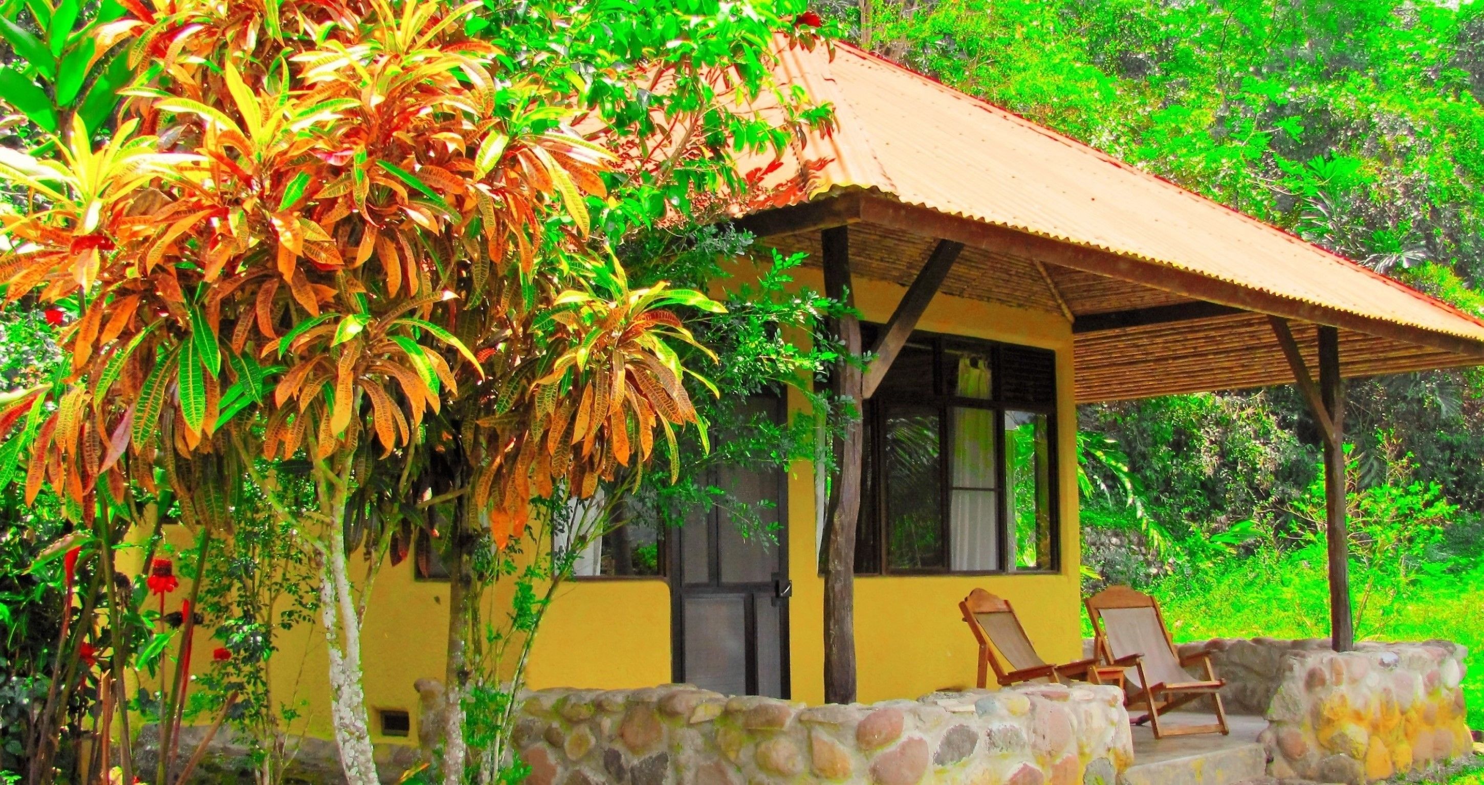 Um pequeno edifício amarelo com telhado de azulejos, uma parede de pedra e duas cadeiras de madeira na frente.