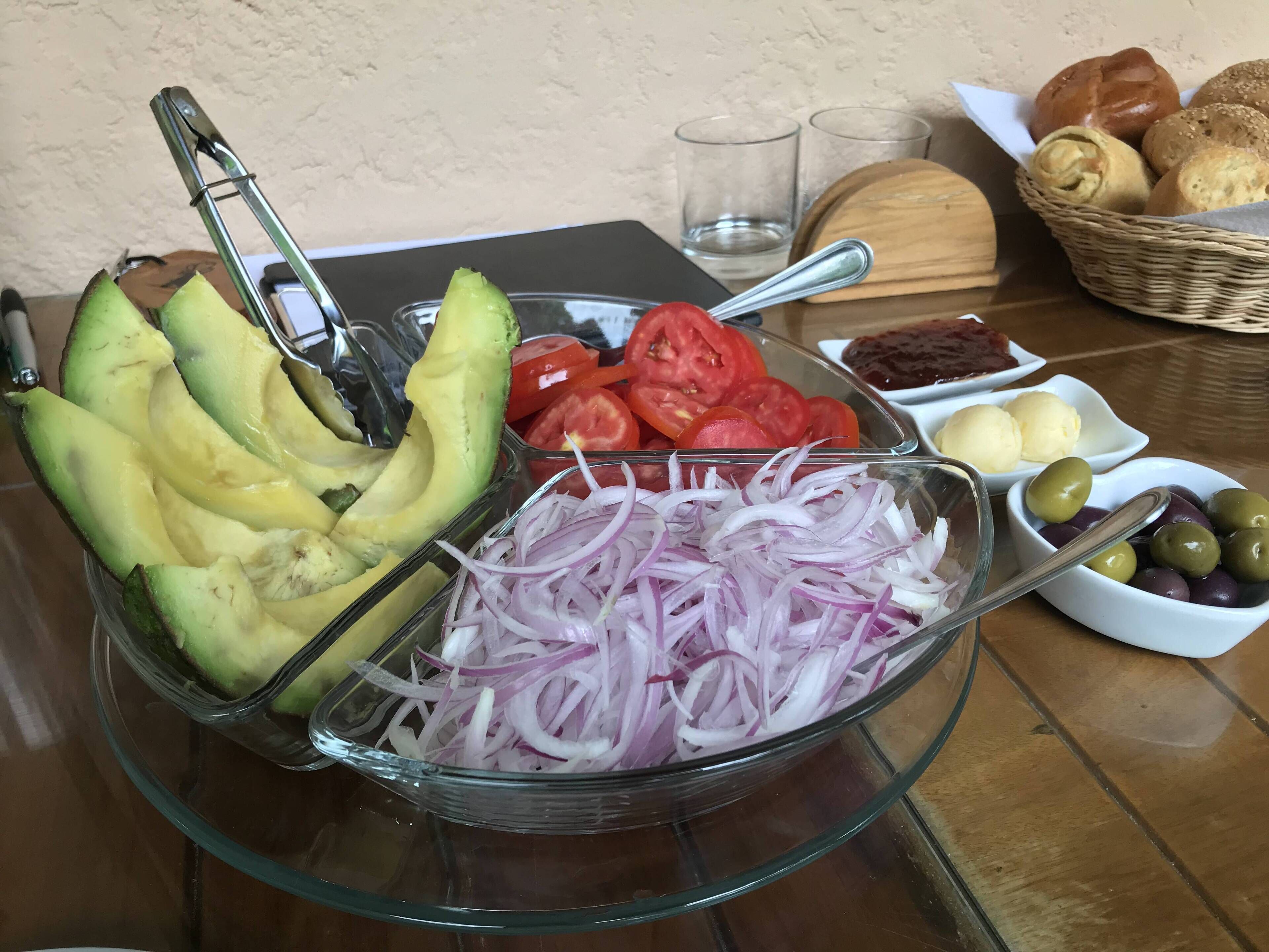 A glass bowl containing sliced avocado, tomato, and red onion on a table.