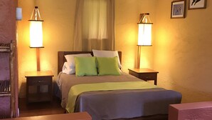 Un dormitorio con cama, dos mesitas de noche, una mesita auxiliar, una ventana con cortinas y dos lámparas de pared.