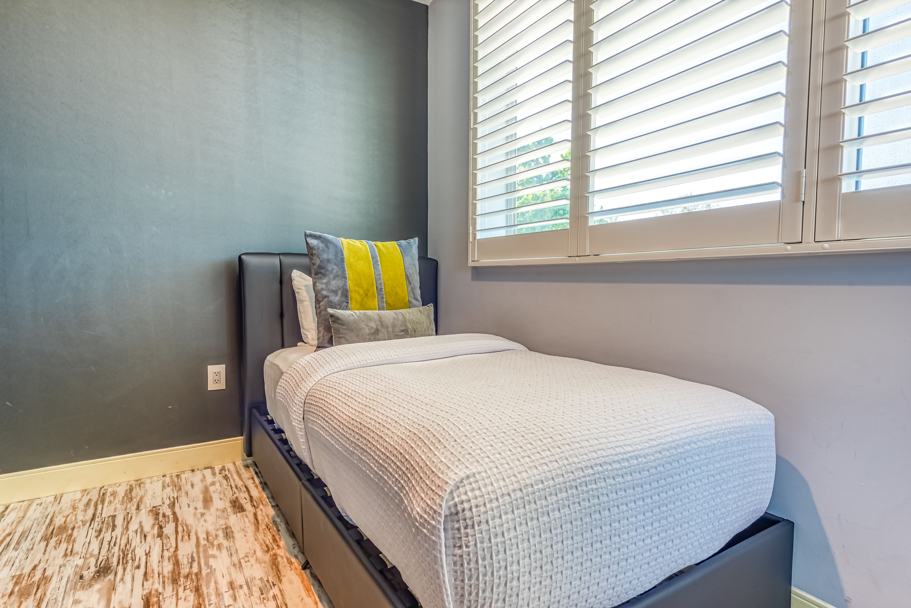 Un dormitorio con cama, almohadas, una ventana con persianas y una alfombra texturizada.