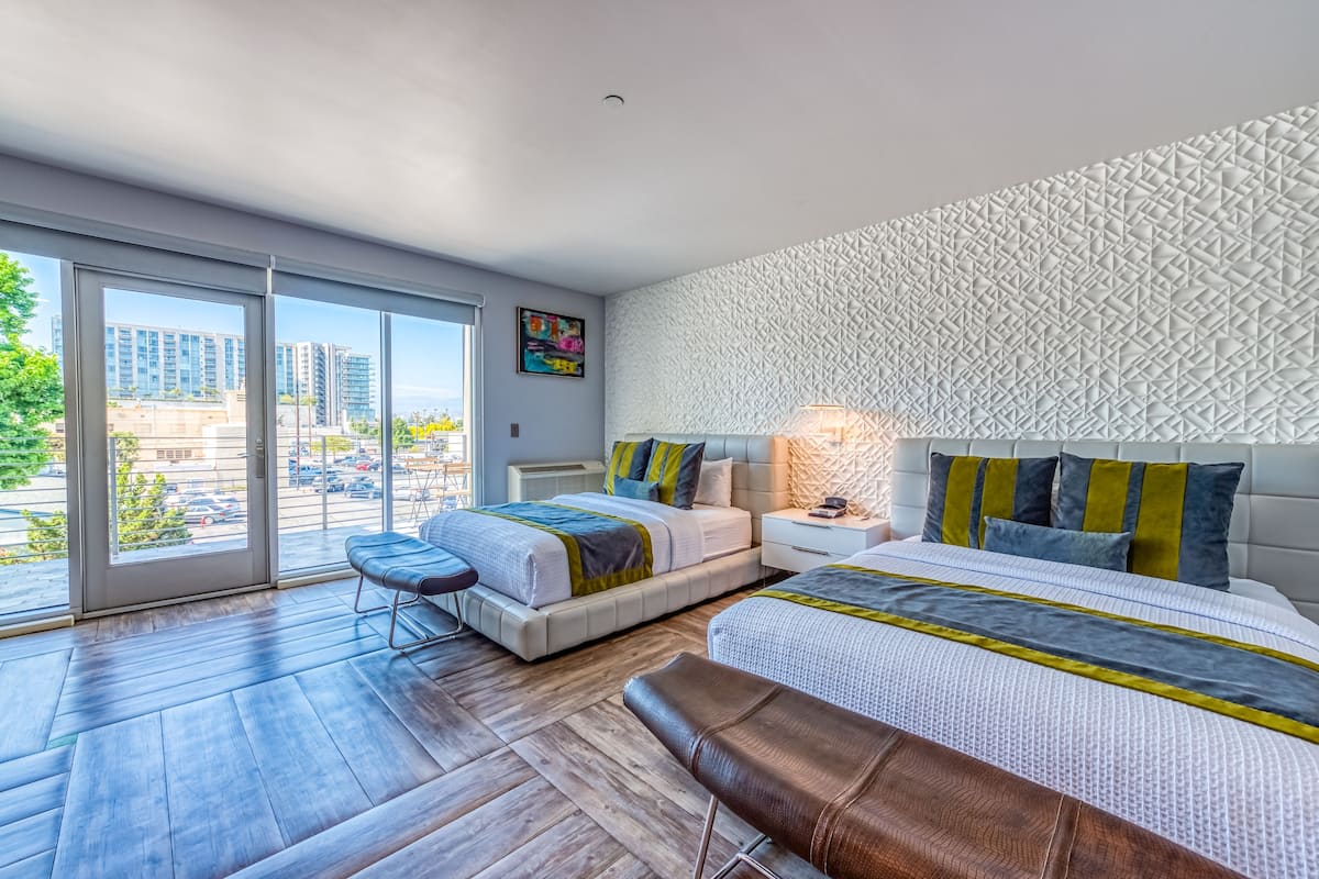 Une chambre d’hôtel moderne dotée d’une grande porte-fenêtre coulissante donnant sur un balcon, d’un lit avec une tête de lit à motifs, d’une table de chevet avec une lampe et d’un banc.
