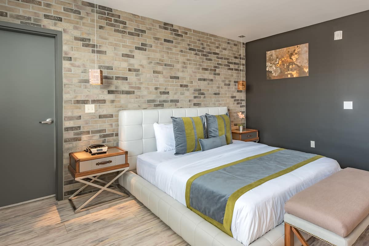 Une chambre moderne avec un mur au motif de briques, une tête de lit blanche recouverte de tissu et un lit agrémenté d’une couvre-lit à rayures grises et vertes.