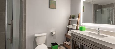 A modern bathroom with a glass shower enclosure, a white toilet, a green trash bin, and a wooden vanity with a mirror.