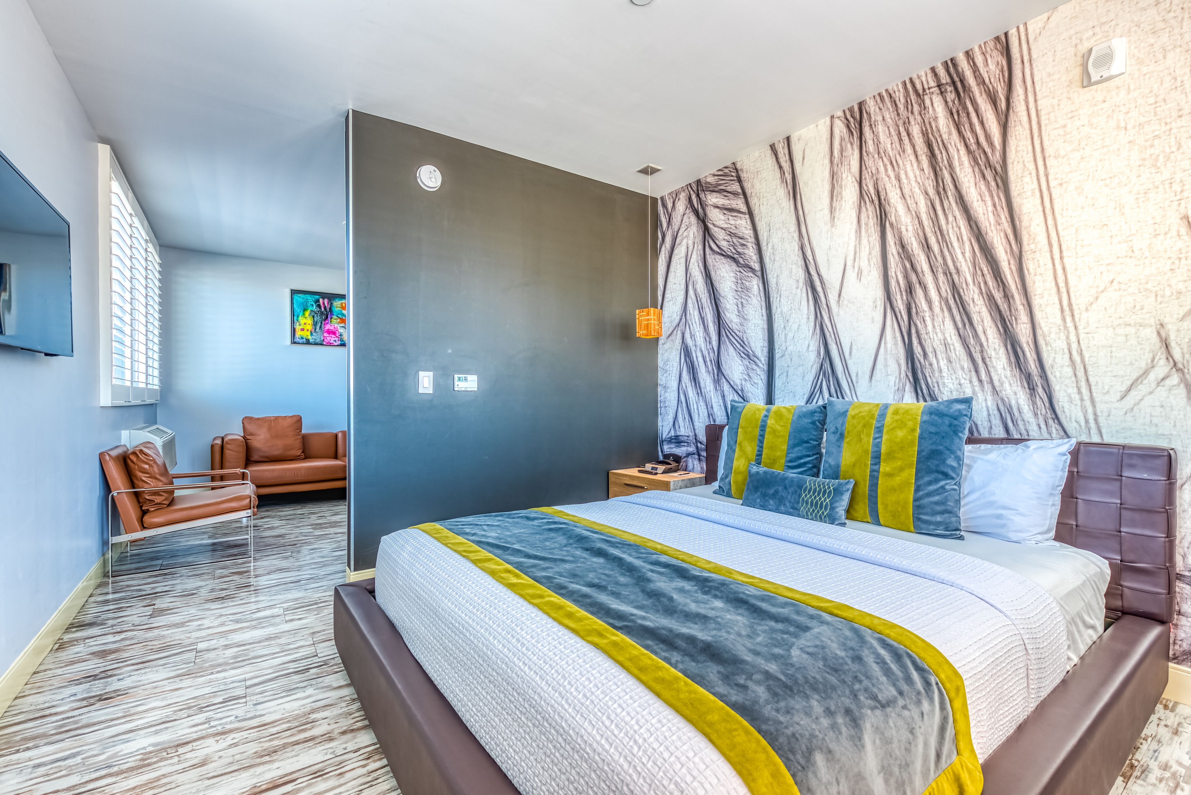 A modern hotel room with a large bed, a seating area with a brown leather sofa, and a large abstract wall mural.