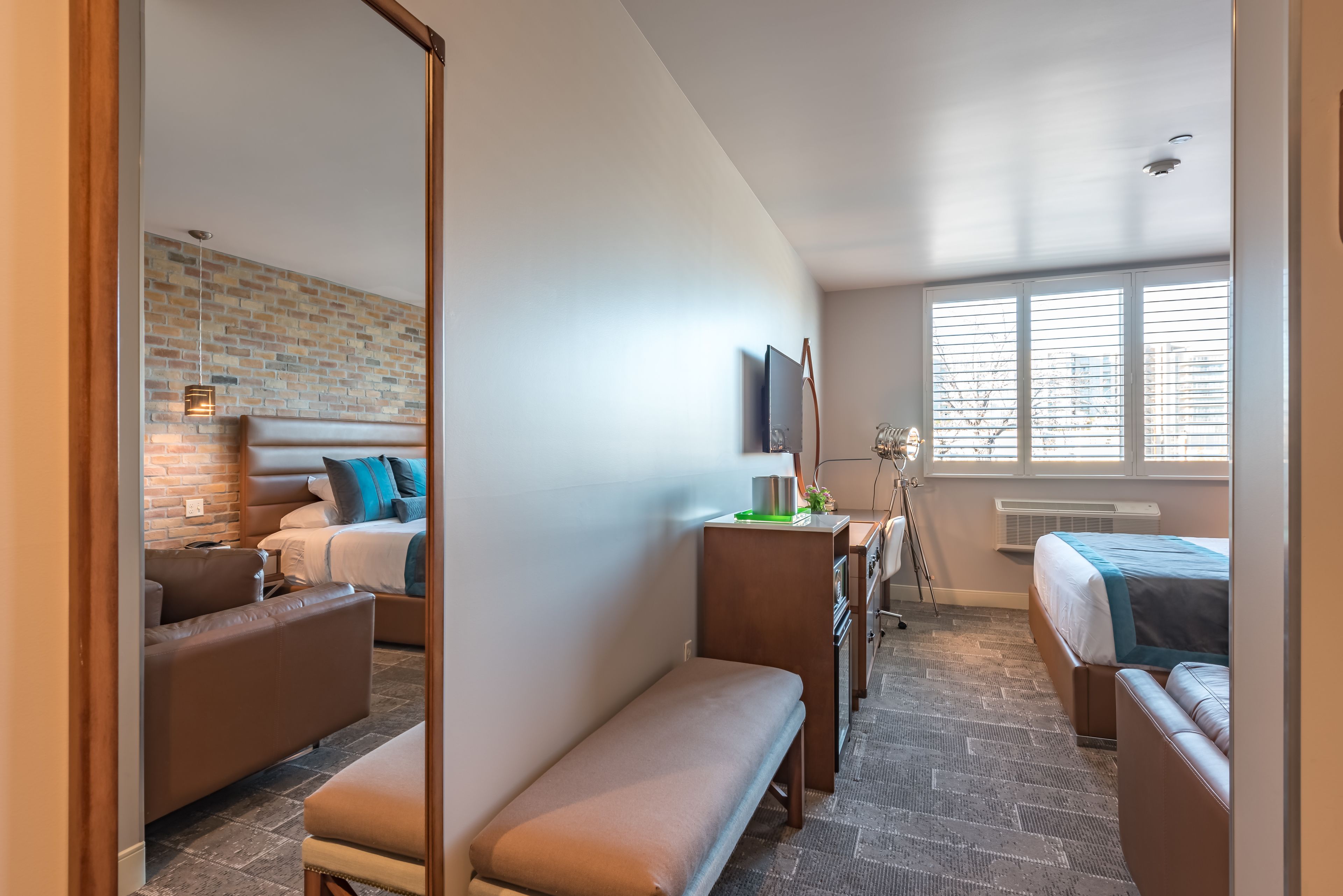 Una habitación de hotel moderna con pared de ladrillo, un ventanal con persianas, una cama con colcha azul y blanca, un sofá de cuero marrón, un banco y un escritorio con lámpara.