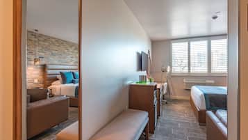 Una habitación de hotel moderna con pared de ladrillo, un ventanal con persianas, una cama con colcha azul y blanca, un sofá de cuero marrón, un banco y un escritorio con lámpara.