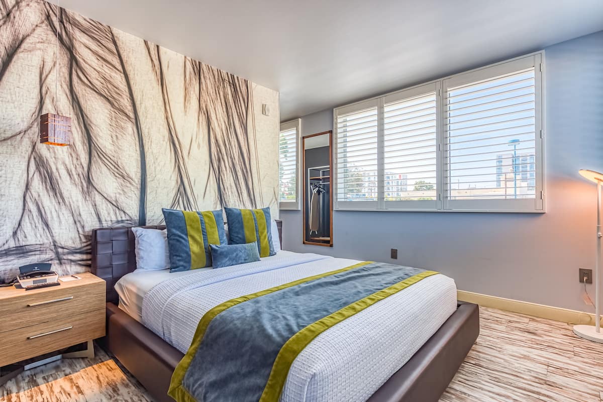 Une chambre à coucher moderne avec un grand lit, une table de chevet, un miroir fixé au mur et une fenêtre avec des stores.