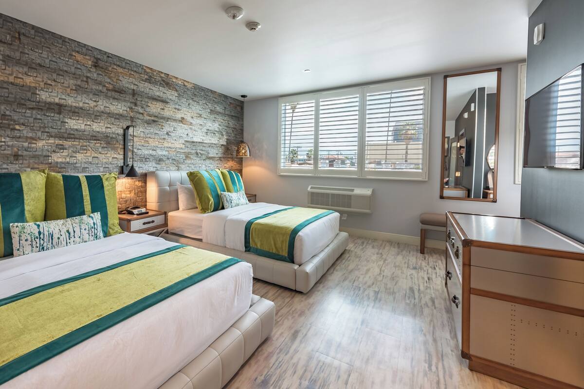 Une chambre moderne avec deux lits, un mur en briques, de grandes fenêtres et une commode.