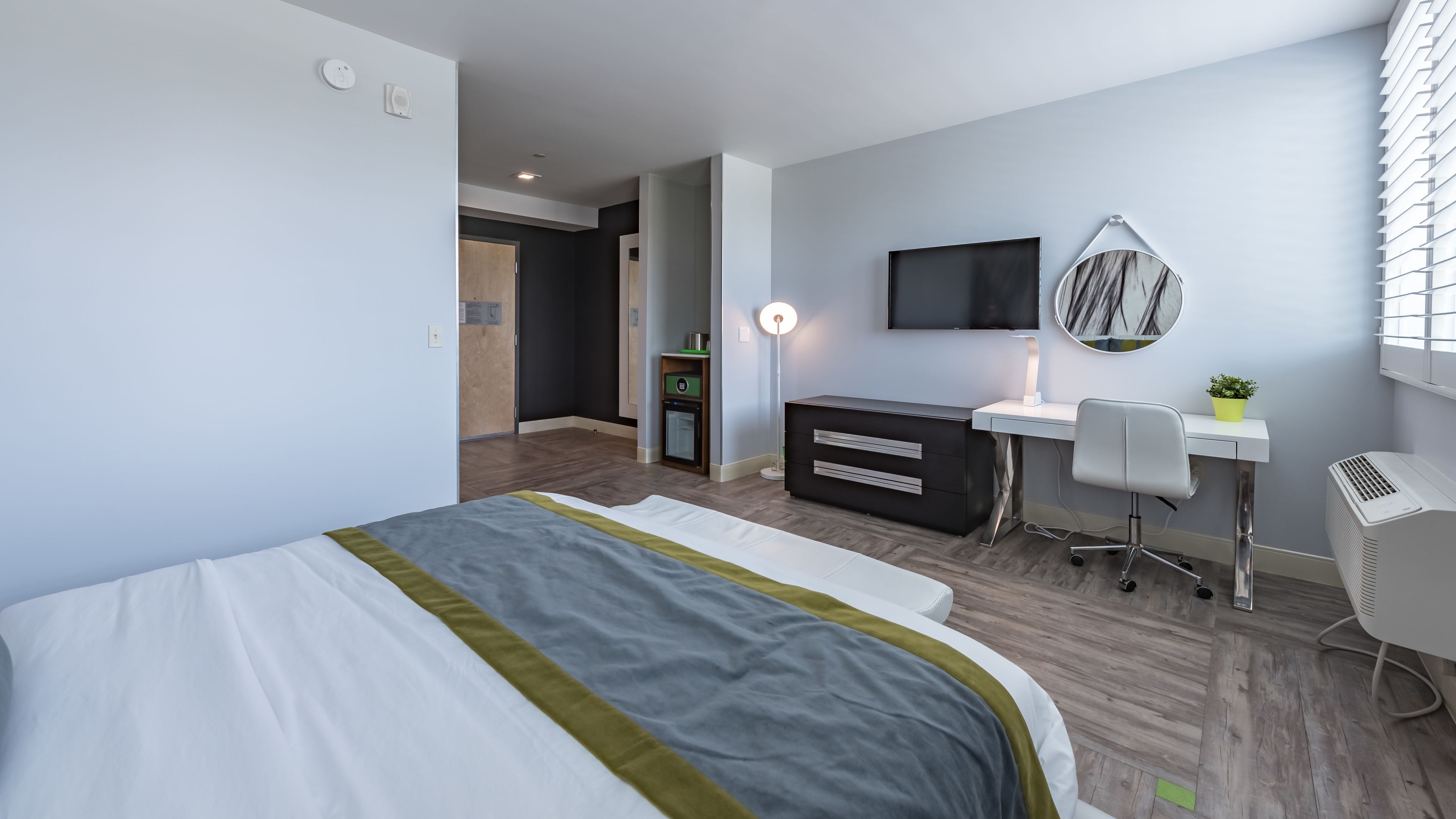 Ett modernt hotellrum med en säng, ett skrivbord, en stol, en tv och en liten växt.
