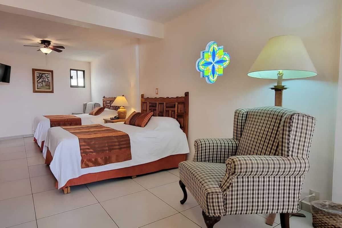 A hotel room with two beds, a chair, a TV, a ceiling fan, and a wall decoration.