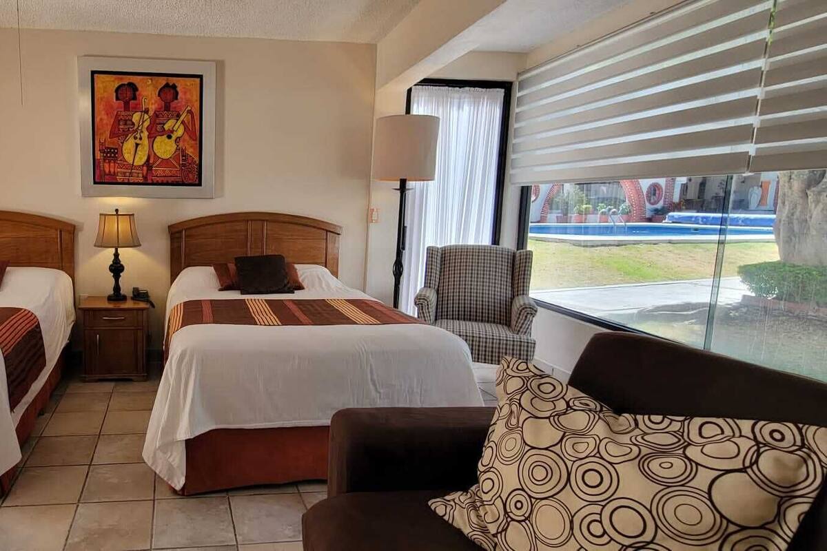 A hotel room with two beds, a sofa, a chair, a lamp, and a window with a view.